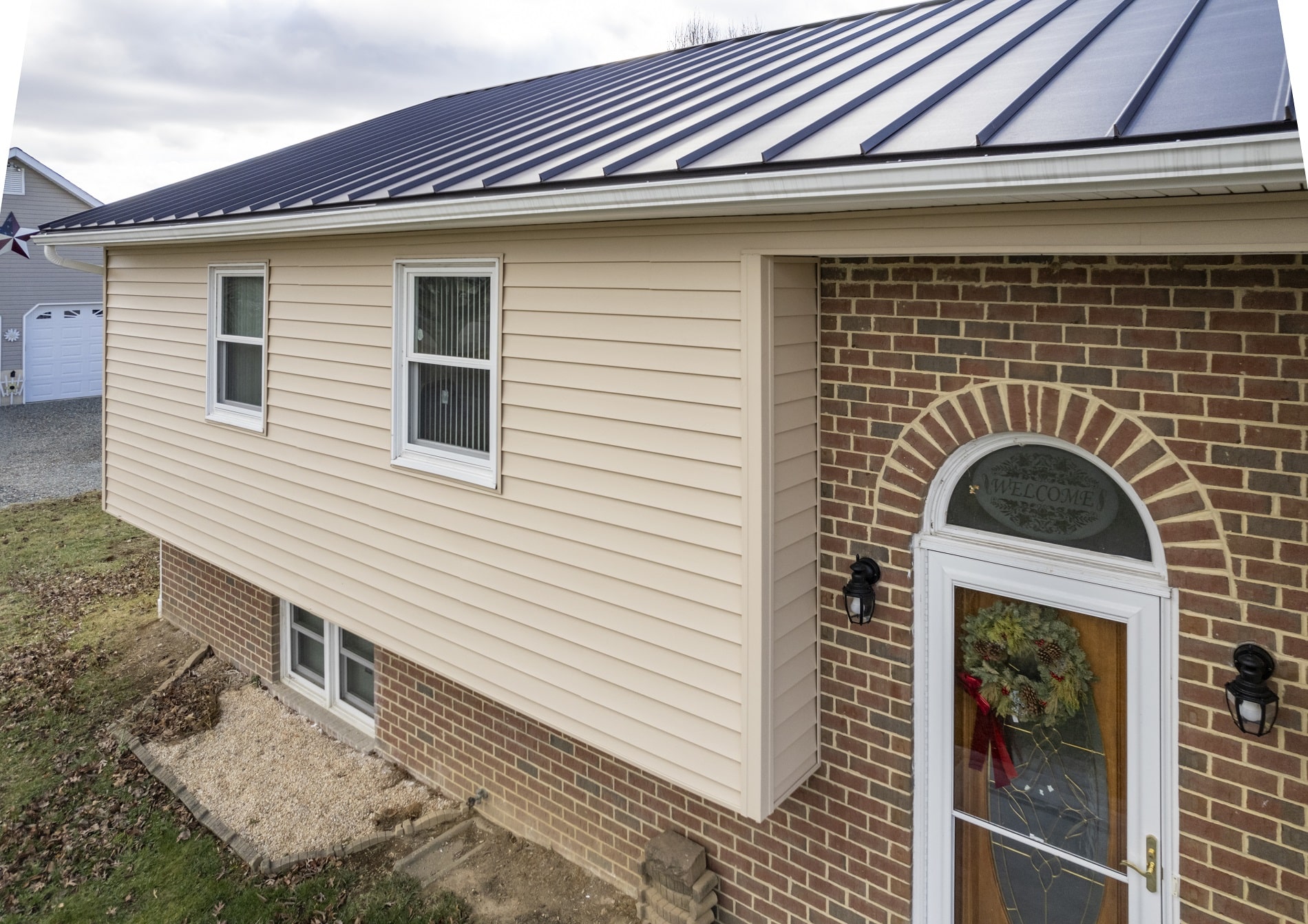 A house with beige siding, brick lower half, and a black metal roof. There are two windows, a glass door with a wreath, and an exterior light fixture.