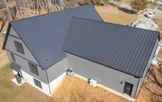 Aerial view of a modern house with a dark gray metal roof and dark gray siding, situated on a sparse lawn with visible HVAC units near the foundation. Aerial view of a modern house with a dark gray metal roof and dark gray siding, situated on a sparse lawn with visible HVAC units near the foundation.