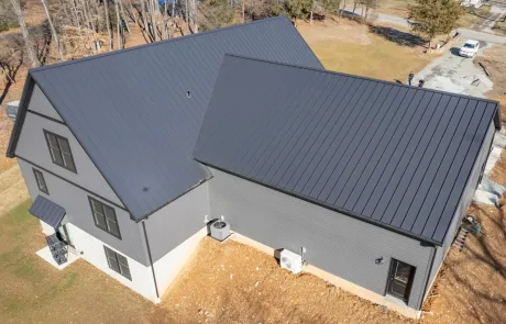 Aerial view of a modern house with a dark gray metal roof and gray siding, situated on a partially grass-covered lot with visible HVAC units next to the building.