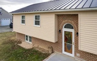 A split-level house with beige siding, brick facade, black metal roof, arched glass front door, and concrete porch. A detached garage is visible in the background. A split-level house with beige siding, brick facade, black metal roof, arched glass front door, and concrete porch. A detached garage is visible in the background.