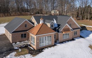 A large brick house with a copper roofed sunroom, surrounded by a snowy lawn and bordered by trees. A large brick house with a copper roofed sunroom, surrounded by a snowy lawn and bordered by trees.