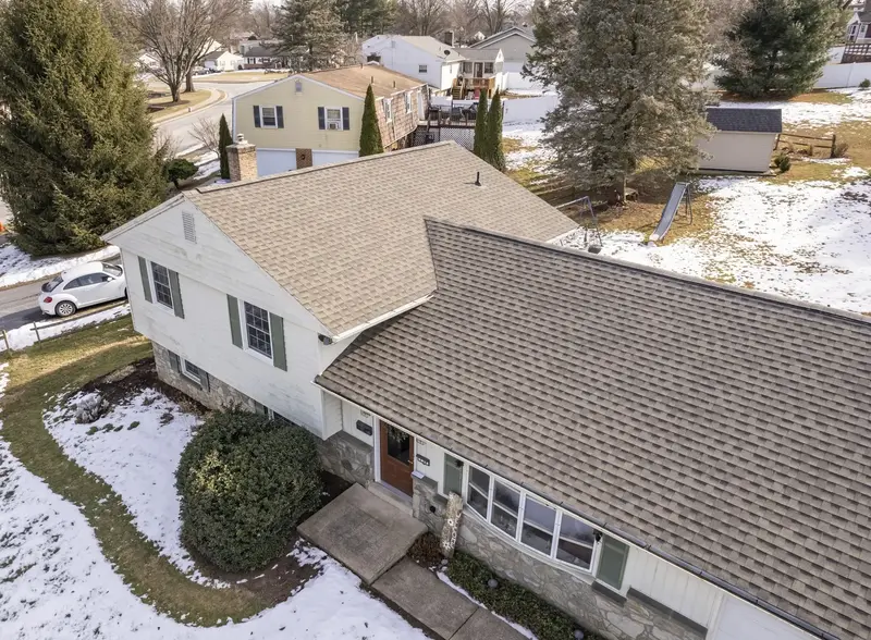 Aerial view of a suburban split-level house with a shingled roof, surrounded by patches of snow, trees, and neighboring homes. Aerial view of a suburban split-level house with a shingled roof, surrounded by patches of snow, trees, and neighboring homes.
