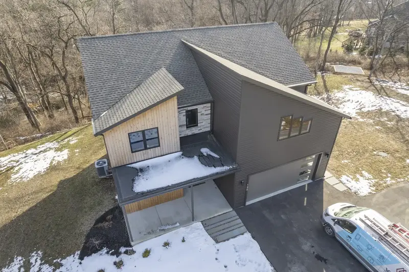 A modern two-story house with mixed wood and dark siding, an attached garage, and a driveway with a work van parked, surrounded by patches of snow and trees.