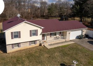 A beige split-level house with a maroon metal roof, attached garage, porch, and a driveway with parked trailer and car; trees and grass in the background. DuraCraft logo in upper left corner.