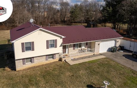 A beige split-level house with a maroon metal roof, attached garage, porch, and a driveway with parked trailer and car; trees and grass in the background. DuraCraft logo in upper left corner.