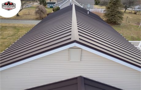 Aerial view of a house roof with brown metal panels and beige siding, surrounded by a yard and trees. DuraCraft logo is visible in the upper left corner.