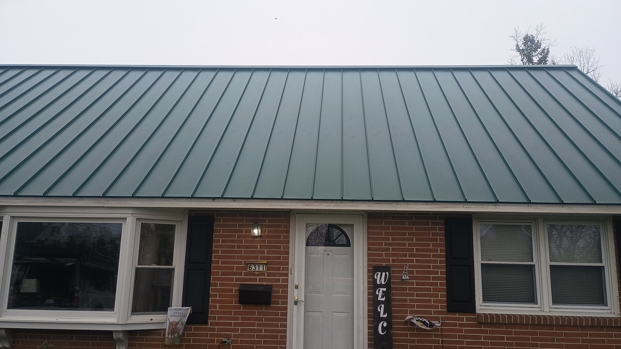 A brick house with a green metal roof, white front door, two black-shuttered windows, and a vertical WELCOME sign by the entrance. A brick house with a green metal roof, white front door, two black-shuttered windows, and a vertical WELCOME sign by the entrance.