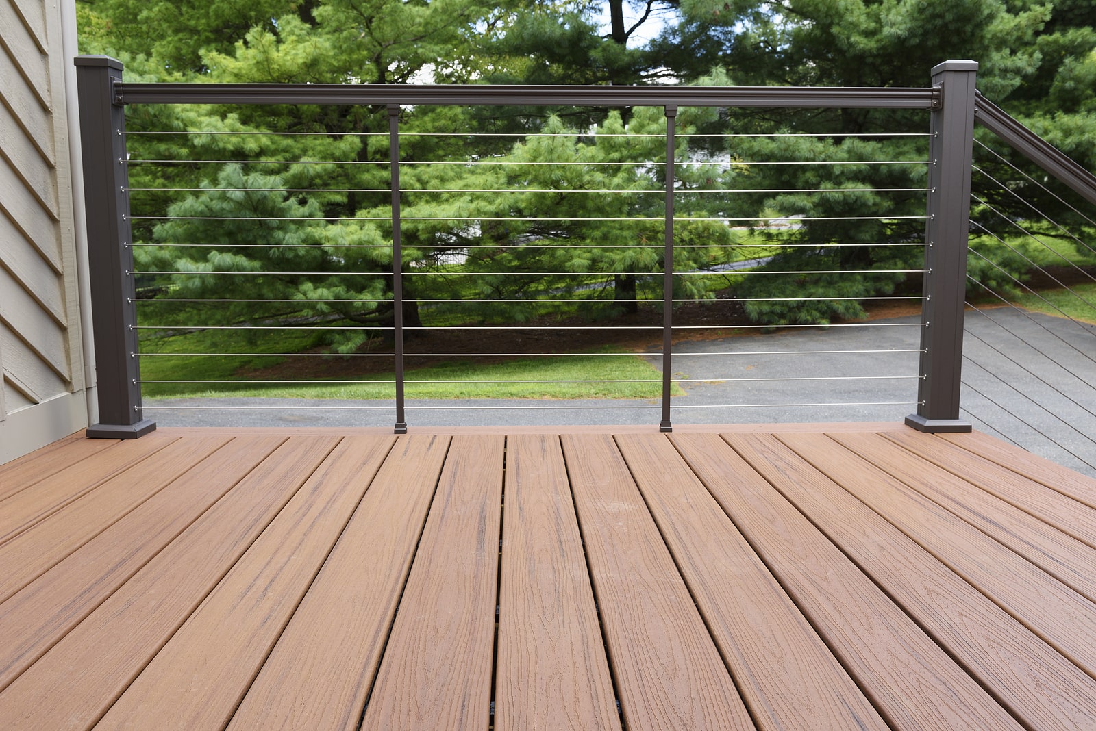 A deck with brown composite wood flooring and a metal cable railing overlooks a driveway and green trees in the background.
