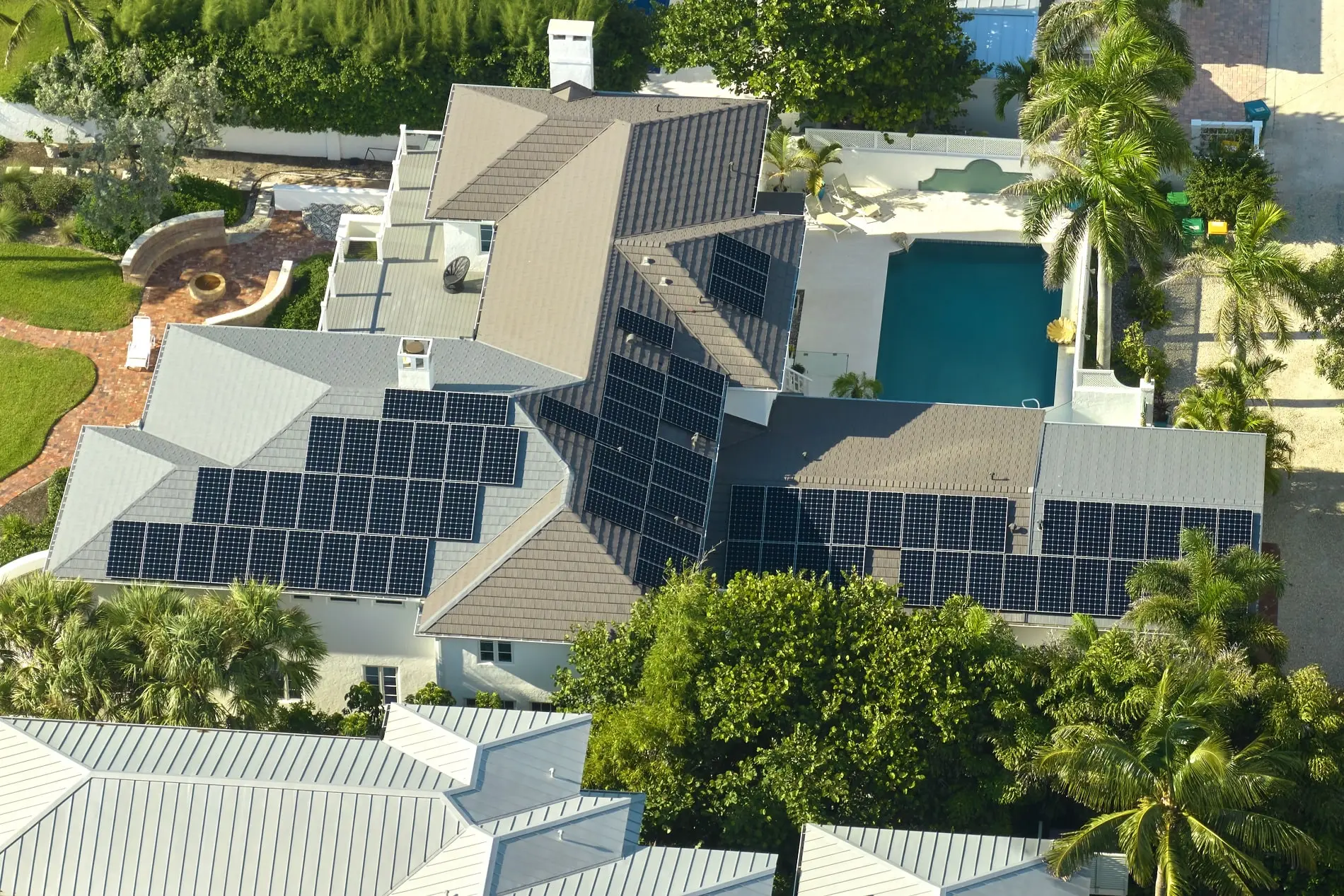 Aerial view of a large house with numerous solar panels installed on the roof, surrounded by trees and featuring a backyard swimming pool. Aerial view of a large house with numerous solar panels installed on the roof, surrounded by trees and featuring a backyard swimming pool.