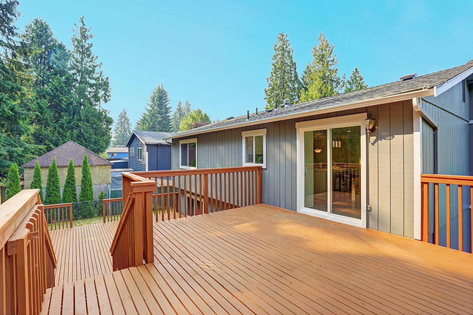 A spacious wooden deck with railing attached to a gray house, surrounded by trees and adjacent houses under a clear blue sky.