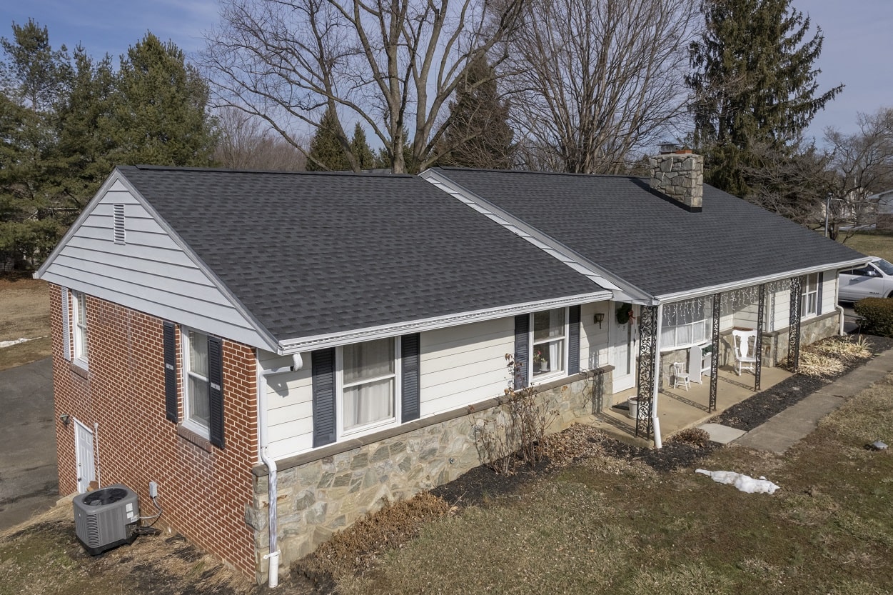 A single-story house with a dark shingle roof, brick and stone exterior, white siding, front porch with chairs, and leafless trees in the background. A single-story house with a dark shingle roof, brick and stone exterior, white siding, front porch with chairs, and leafless trees in the background.