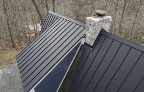 A metal roof with standing seam panels and a stone chimney, surrounded by leafless trees in a wooded area.
